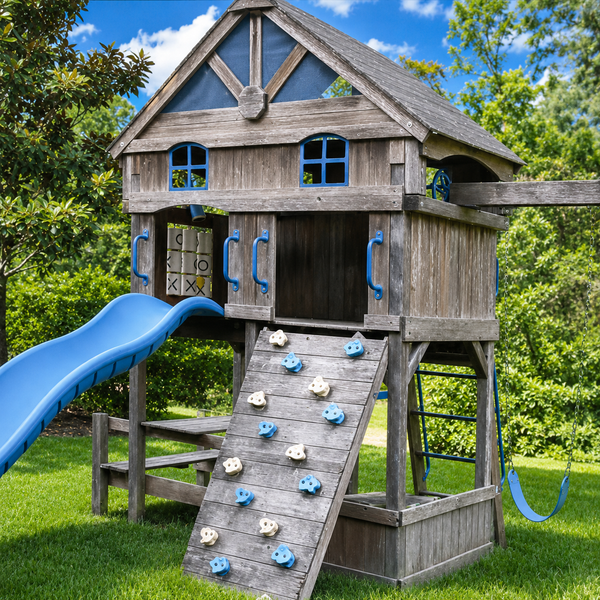 A large wooden playhouse with a gray-brown weathered wood finish, featuring a blue slide, climbing wall, windows, and blue handles. It is set in a backyard with green grass and trees under a blue sky.
